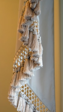 A close-up view of a textile with intricate macramé patterns and numerous fringed tassels in neutral tones. The woven design features a combination of tightly wrapped and loosely knotted textures, creating a sense of handcrafted elegance.