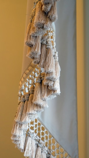 A close-up view of a textile with intricate macramé patterns and numerous fringed tassels in neutral tones. The woven design features a combination of tightly wrapped and loosely knotted textures, creating a sense of handcrafted elegance.