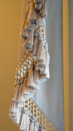 A close-up view of a textile with intricate macram&eacute; patterns and numerous fringed tassels in neutral tones. The woven design features a combination of tightly wrapped and loosely knotted textures, creating a sense of handcrafted elegance.