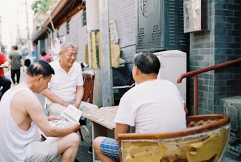A group of elderly men are sitting in chairs outdoors, engaged in a game on a small table. One man holds a newspaper while another is focused on the game pieces. They are seated in an alleyway beside a building with a tiled wall.