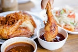 A hand dipping a crispy fried chicken drumstick into a bowl of reddish-brown sauce. On the table are additional pieces of fried chicken on a plate and a salad consisting of vegetables and possibly some meat.