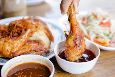 A hand dipping a crispy fried chicken drumstick into a bowl of reddish-brown sauce. On the table are additional pieces of fried chicken on a plate and a salad consisting of vegetables and possibly some meat.