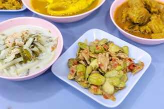 A selection of colorful dishes displayed on a table. The foreground features a dish with sliced vegetables and pieces of meat, accompanied by a sauce. Another dish appears to contain a creamy soup with leafy greens and prawns. In the background, there are two more dishes, one with vibrant yellow curry and another with a darker, spiced sauce.