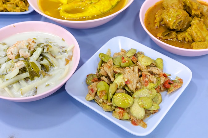 A selection of colorful dishes displayed on a table. The foreground features a dish with sliced vegetables and pieces of meat, accompanied by a sauce. Another dish appears to contain a creamy soup with leafy greens and prawns. In the background, there are two more dishes, one with vibrant yellow curry and another with a darker, spiced sauce.