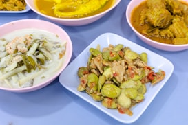 A selection of colorful dishes displayed on a table. The foreground features a dish with sliced vegetables and pieces of meat, accompanied by a sauce. Another dish appears to contain a creamy soup with leafy greens and prawns. In the background, there are two more dishes, one with vibrant yellow curry and another with a darker, spiced sauce.