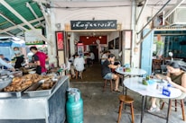 Inside a small restaurant, people are dining at simple round tables. The focus is on the cooking area with trays of food in the foreground. Several individuals, including staff in aprons and customers, are present. The restaurant has a rustic, casual atmosphere with open walls and a green corrugated roof.