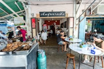 Inside a small restaurant, people are dining at simple round tables. The focus is on the cooking area with trays of food in the foreground. Several individuals, including staff in aprons and customers, are present. The restaurant has a rustic, casual atmosphere with open walls and a green corrugated roof.