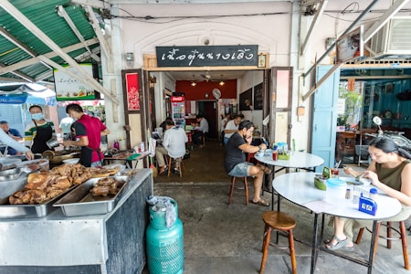 Inside a small restaurant, people are dining at simple round tables. The focus is on the cooking area with trays of food in the foreground. Several individuals, including staff in aprons and customers, are present. The restaurant has a rustic, casual atmosphere with open walls and a green corrugated roof.