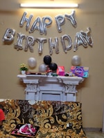 Silver balloons spell out 'Happy Birthday' against a beige wall. Below, a white mantelpiece is adorned with various decorative items including flowers and gift boxes. Three balloons, one white and two black, are positioned among the decorations. A couch with an ornate floral design covers part of the image, with a tray of rose petals placed on it.