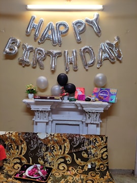 Silver balloons spell out 'Happy Birthday' against a beige wall. Below, a white mantelpiece is adorned with various decorative items including flowers and gift boxes. Three balloons, one white and two black, are positioned among the decorations. A couch with an ornate floral design covers part of the image, with a tray of rose petals placed on it.