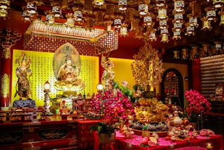 An ornately decorated shrine filled with intricate golden statues, richly detailed carvings, and colorful floral arrangements. The statues display various deities or figures, surrounded by vibrant flowers and lit by elaborate hanging lanterns. The background features a wall covered in traditional script, contributing to the spiritual and cultural ambiance.