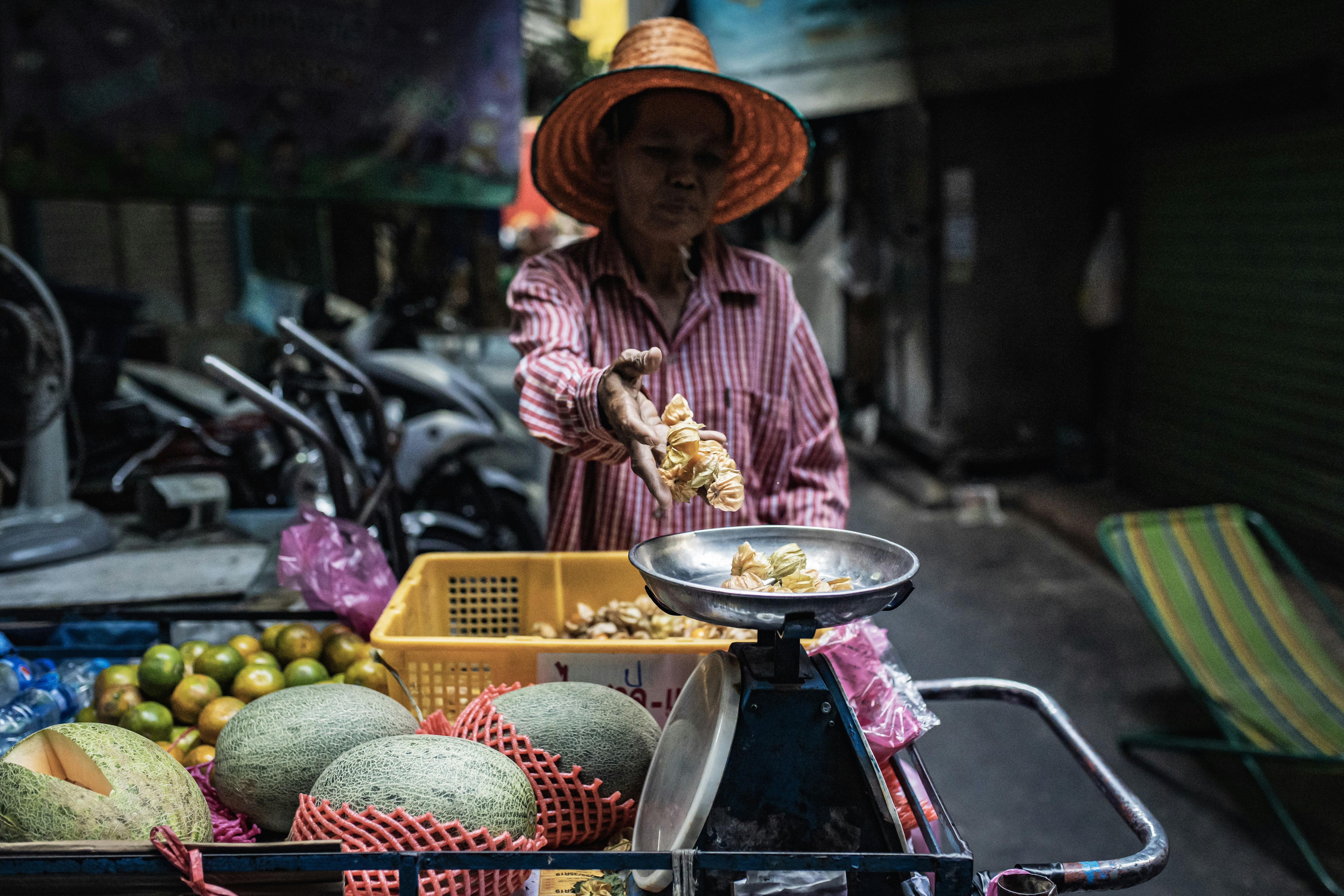 man in red and white stripe dress shirt and brown hat holding brown wooden tray with near near near on, Fruit seller in Bangkok