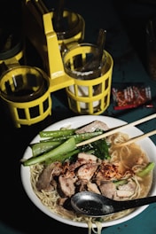 A bowl filled with a noodle soup garnished with slices of meat, leafy green vegetables, and a sprinkling of chili flakes. The bowl is accompanied by a metal spoon and two chopsticks resting on the rim. In the background, there is a yellow condiment caddy holding several containers with metal spoons.