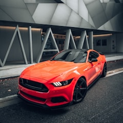 A sleek, modern sports car is parked on a city street. The car is bright orange with a glossy finish, contrasting against the urban backdrop. The vehicle's design features sharp, angular lines and black wheels, while its headlights are uniquely styled. The background showcases a geometric architectural structure with large, grey panels and the word 'MIAMI' in bold letters.