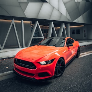 A sleek, modern sports car is parked on a city street. The car is bright orange with a glossy finish, contrasting against the urban backdrop. The vehicle's design features sharp, angular lines and black wheels, while its headlights are uniquely styled. The background showcases a geometric architectural structure with large, grey panels and the word 'MIAMI' in bold letters.