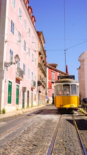 A classic yellow tram travels down a cobblestone street lined with pastel-colored buildings under a clear blue sky. The architecture is traditional with wrought iron balconies and large windows, suggesting a historic European city atmosphere.