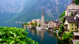 A picturesque lakeside town featuring traditional European architecture. A prominent church with a tall spire is situated near the water's edge, surrounded by colorful buildings. The serene lake reflects the structures, and lush green mountains provide a stunning backdrop. Foreground plants add vibrant greenery to the scene.