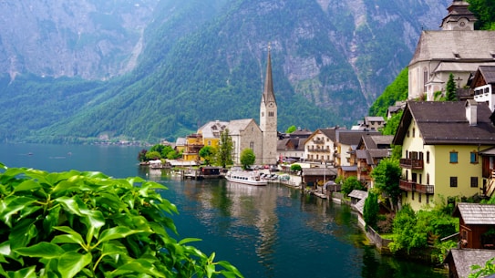 A picturesque lakeside town featuring traditional European architecture. A prominent church with a tall spire is situated near the water's edge, surrounded by colorful buildings. The serene lake reflects the structures, and lush green mountains provide a stunning backdrop. Foreground plants add vibrant greenery to the scene.