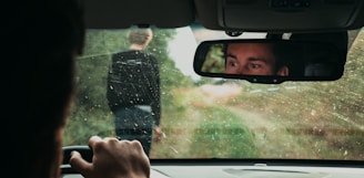 A person is sitting inside a car, gripping the steering wheel, looking at the rearview mirror. Another person is seen through the windshield, walking on a dirt road surrounded by greenery. The windshield is slightly dirty, and the driver appears to be focused or concerned.