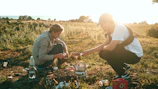 Two people are cooking outdoors on a grassy field during sunset. They are using a portable stove and have various cooking items scattered around. One is handing a pot to the other, who is crouched on the ground. The background shows a serene landscape with trees and open space. The scene is lit with warm sunset light.