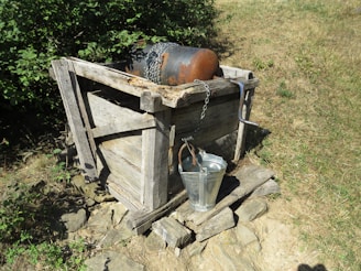 A rustic wooden structure with a weathered appearance is situated outdoors. It holds a large metal cylinder secured by a chain. In front of it, a shiny metal bucket is attached, resting on uneven wooden planks atop a stone base. Surrounding the area, patches of grass and green foliage are visible.