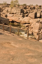 A rocky landscape features large boulders surrounding a small stream of water. The boulders create various shapes and formations, with a few scattered trees in the background. A metal railing runs along the foreground, separating the viewer from the rocky terrain.
