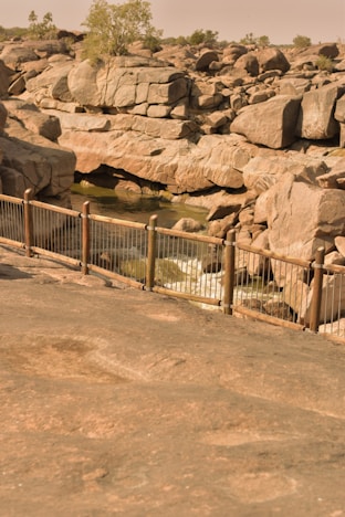 A rocky landscape features large boulders surrounding a small stream of water. The boulders create various shapes and formations, with a few scattered trees in the background. A metal railing runs along the foreground, separating the viewer from the rocky terrain.