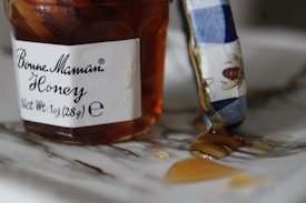 A jar of honey with a label that reads 'Bonne Maman Honey' and a net weight of 1oz (28g) is partially open. Some honey has dripped onto the surface, and there is a blue and white utensil, possibly a butter knife, with a bee design on it, partially covered in honey.