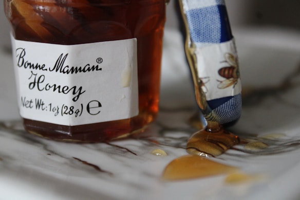 A jar of honey with a label that reads 'Bonne Maman Honey' and a net weight of 1oz (28g) is partially open. Some honey has dripped onto the surface, and there is a blue and white utensil, possibly a butter knife, with a bee design on it, partially covered in honey.