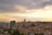 Urban landscape with numerous residential and commercial buildings spread across a hilly area. Tall apartment complexes and mid-rise buildings dominate the skyline. The scene is set against a backdrop of a cloudy sky with hues of orange and yellow, suggesting a sunset or sunrise.