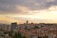 Urban landscape with numerous residential and commercial buildings spread across a hilly area. Tall apartment complexes and mid-rise buildings dominate the skyline. The scene is set against a backdrop of a cloudy sky with hues of orange and yellow, suggesting a sunset or sunrise.