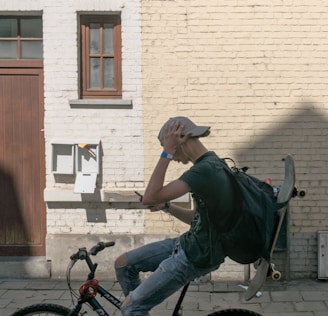 A teenage boy wearing a cap is riding a bicycle while holding his smartphone. A skateboard is strapped to his backpack. He is positioned against a light-colored brick wall with small windows and a mailbox. He appears to be listening to music through earphones.