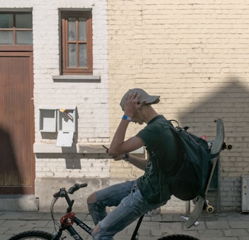 A teenage boy wearing a cap is riding a bicycle while holding his smartphone. A skateboard is strapped to his backpack. He is positioned against a light-colored brick wall with small windows and a mailbox. He appears to be listening to music through earphones.
