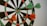 A dartboard with four darts embedded, two green and two orange, indicating an active game or practice session. The darts are positioned close to the center, suggesting accuracy and skill.