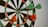 A dartboard with four darts embedded, two green and two orange, indicating an active game or practice session. The darts are positioned close to the center, suggesting accuracy and skill.