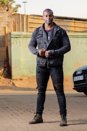 A person stands confidently in an urban setting, wearing a black denim jacket, ripped black jeans, and brown boots. The background features a distressed wall and parts of a parked car.