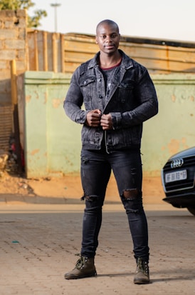 A person stands confidently in an urban setting, wearing a black denim jacket, ripped black jeans, and brown boots. The background features a distressed wall and parts of a parked car.