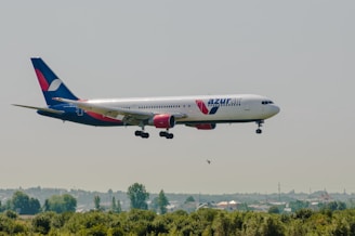 An Azur Air passenger airplane with red and blue accents is flying low over a landscape with green vegetation and hills in the background.