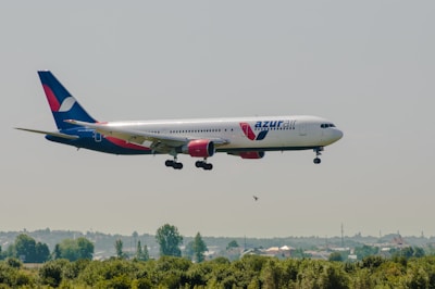 An Azur Air passenger airplane with red and blue accents is flying low over a landscape with green vegetation and hills in the background.
