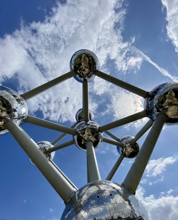 A large architectural structure consists of interconnected metallic spheres, with shiny surfaces reflecting the sky and clouds. The design resembles a molecular structure, towering against a backdrop of a clear blue sky with scattered white clouds.