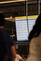 A digital airport departures board displays various flight details including times, destinations, flight numbers, and statuses. The background includes blurred figures of people, suggesting movement and activity. Overhead lighting and industrial design elements are visible, hinting at an indoor airport setting.