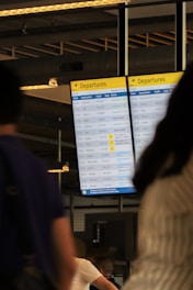 A digital airport departures board displays various flight details including times, destinations, flight numbers, and statuses. The background includes blurred figures of people, suggesting movement and activity. Overhead lighting and industrial design elements are visible, hinting at an indoor airport setting.