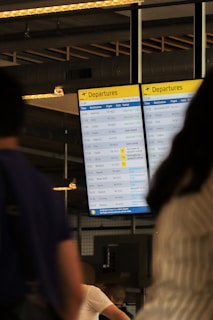 A digital airport departures board displays various flight details including times, destinations, flight numbers, and statuses. The background includes blurred figures of people, suggesting movement and activity. Overhead lighting and industrial design elements are visible, hinting at an indoor airport setting.