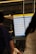 A digital airport departures board displays various flight details including times, destinations, flight numbers, and statuses. The background includes blurred figures of people, suggesting movement and activity. Overhead lighting and industrial design elements are visible, hinting at an indoor airport setting.