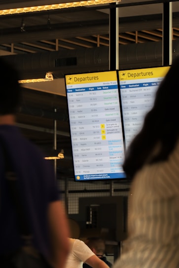 A digital airport departures board displays various flight details including times, destinations, flight numbers, and statuses. The background includes blurred figures of people, suggesting movement and activity. Overhead lighting and industrial design elements are visible, hinting at an indoor airport setting.
