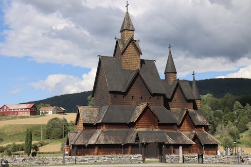 A large wooden church with distinctive steeply pitched roofs, located in a rural setting. The church features intricate architectural details and multiple spires topped with crosses. Surrounding the church is a lush landscape with trees, fields, and a few buildings in the background under a partly cloudy sky.
