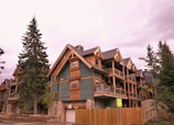 Wooden multi-story chalets with green paneling and stone foundations, surrounded by tall evergreen trees under an overcast sky. A 'School Zone Ends' sign is visible on a wooden fence nearby.