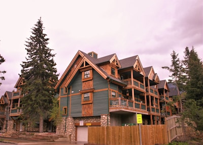 Wooden multi-story chalets with green paneling and stone foundations, surrounded by tall evergreen trees under an overcast sky. A 'School Zone Ends' sign is visible on a wooden fence nearby.