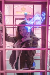 A person is standing inside a vintage-style pink telephone booth, holding a phone receiver to their ear. There is a warm, ambient pink lighting throughout the scene, with neon blue light reflections on the glass. The person is wearing a brown shearling coat and has long hair.