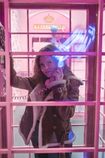A person is standing inside a vintage-style pink telephone booth, holding a phone receiver to their ear. There is a warm, ambient pink lighting throughout the scene, with neon blue light reflections on the glass. The person is wearing a brown shearling coat and has long hair.